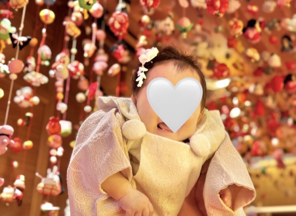 つるし雛を背景に、明るい照明の下で赤ちゃんを高く抱き上げて撮影したひな祭りフォト。壁一面に可愛い布細工が並ぶ