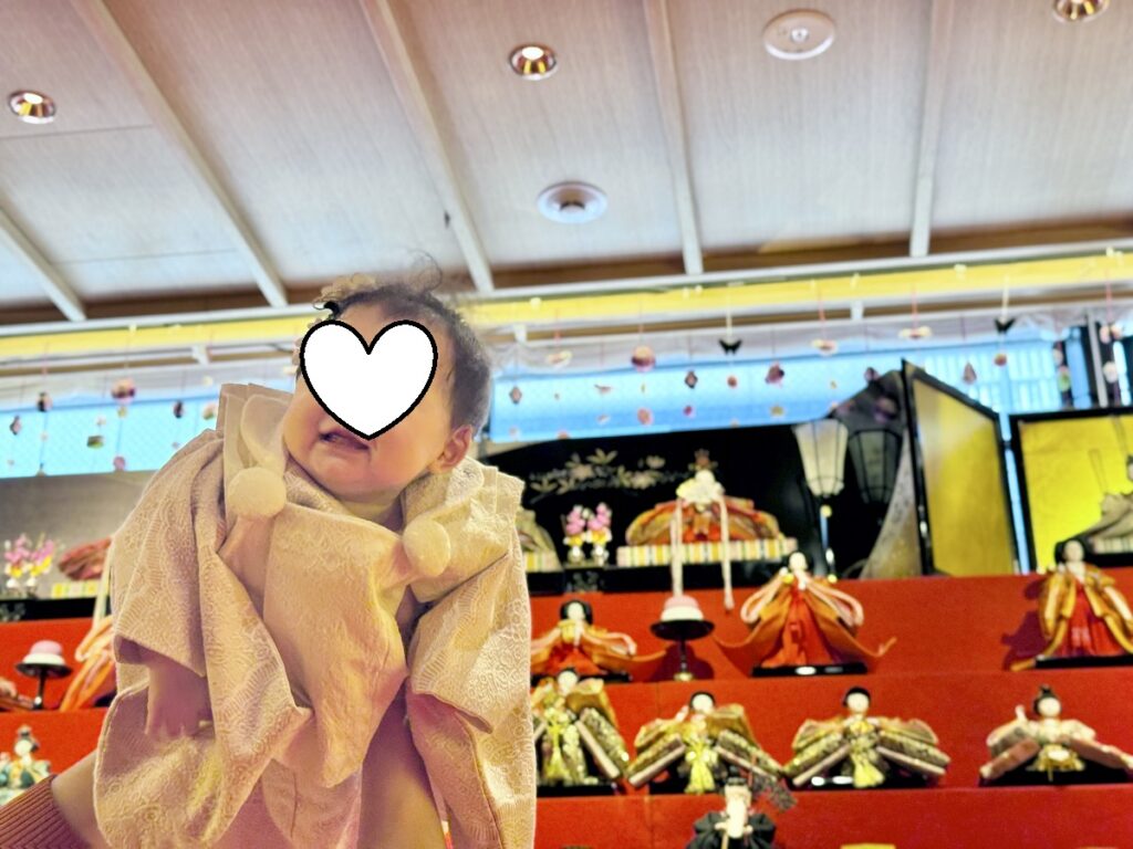 雛壇の前で赤ちゃんを抱き上げて撮影した近景。段飾りの人形や装飾が背景に広がり、ひな祭りらしい雰囲気が伝わる