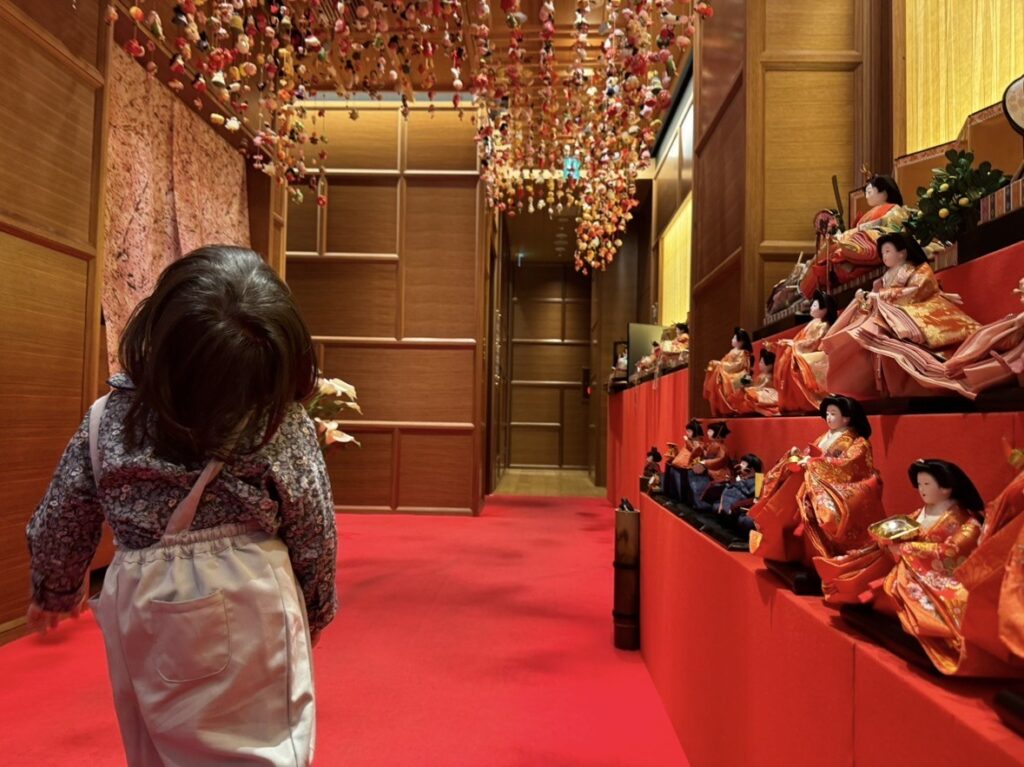 赤い絨毯の通路に面して雛人形が並ぶ展示。頭上にはつるし雛が連なり、子どもが展示方向へ歩いていく様子