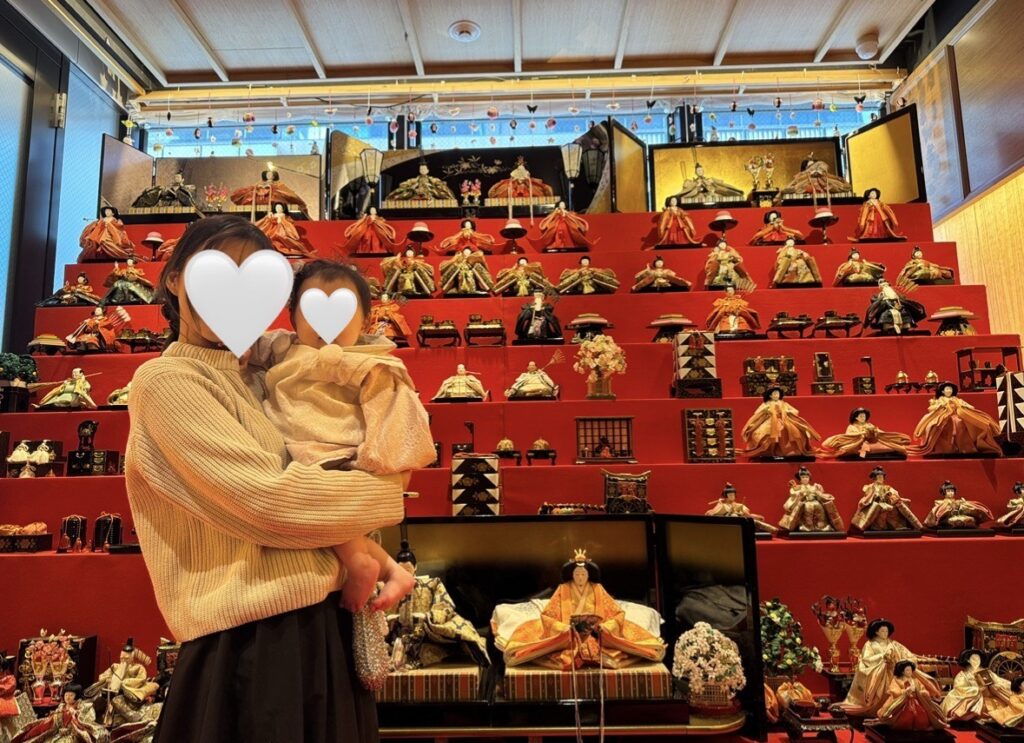 赤い段飾りの雛人形を背に、親子で記念撮影している様子。屏風や装飾が整然と並ぶ華やかな雛壇展示